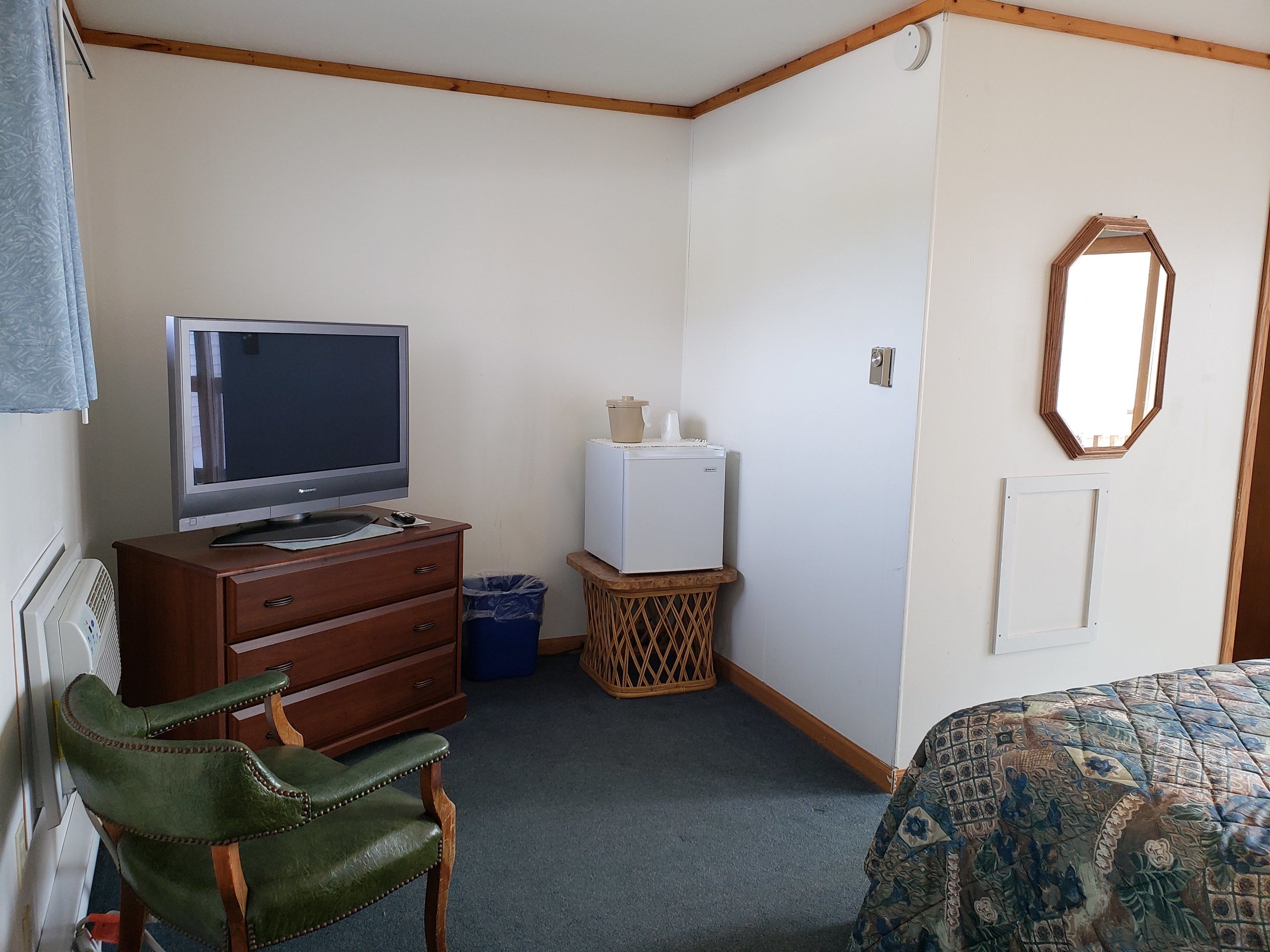 A small, simple room with a television, a small refrigerator, a wooden cabinet, a wicker basket, a green leather chair, and a patterned bedspread.