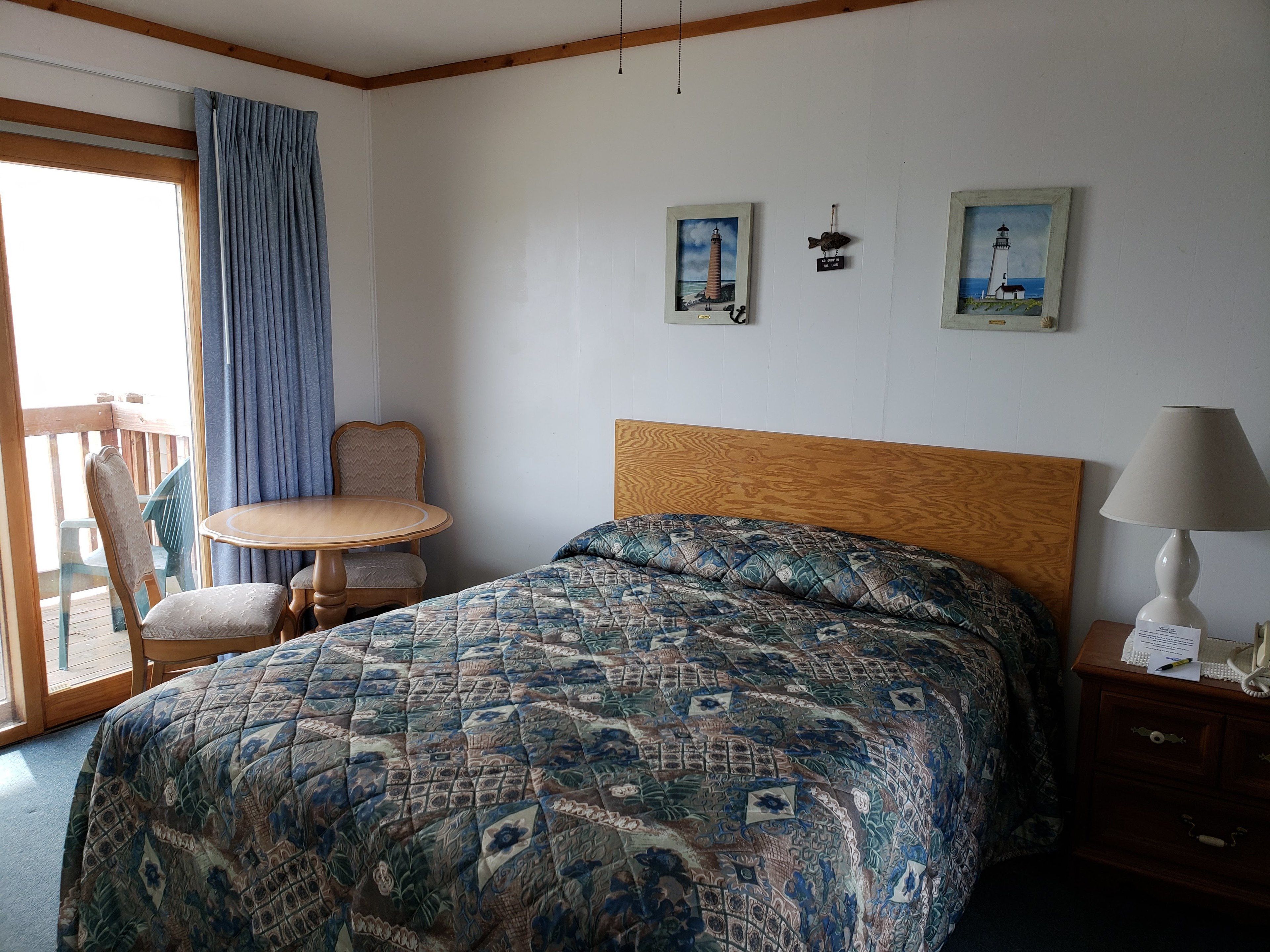 Een slaapkamer met een bed, een houten hoofdbord, een nachtkastje met een lamp, een stoel, een tafel en twee ingekaderde foto’s aan de muur.