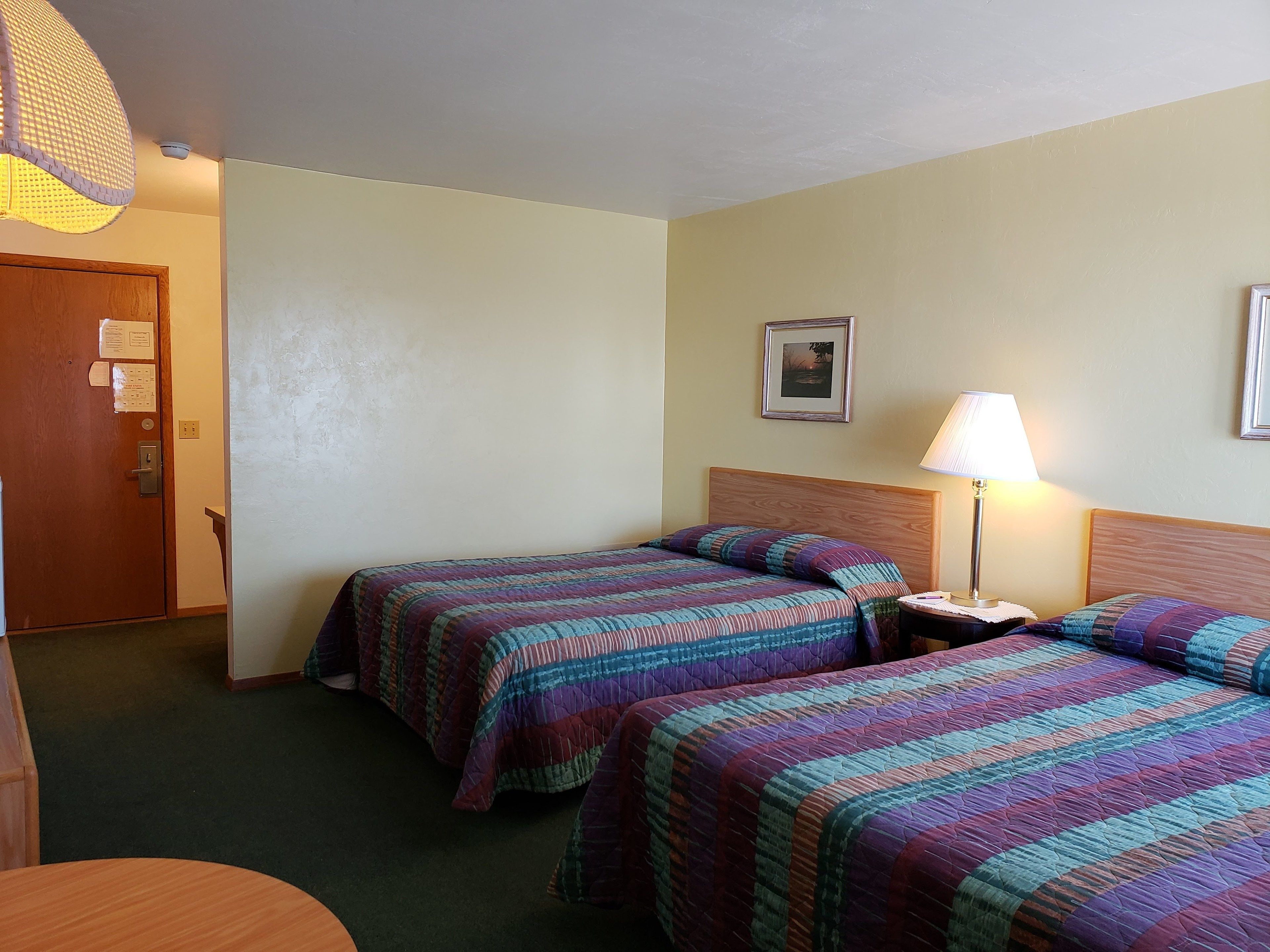 Ein Hotelzimmer mit zwei Betten, einem hölzernen Kopfteil, einem Nachttisch mit Lampe, einer Tür mit Schloss und einem Wandbild.