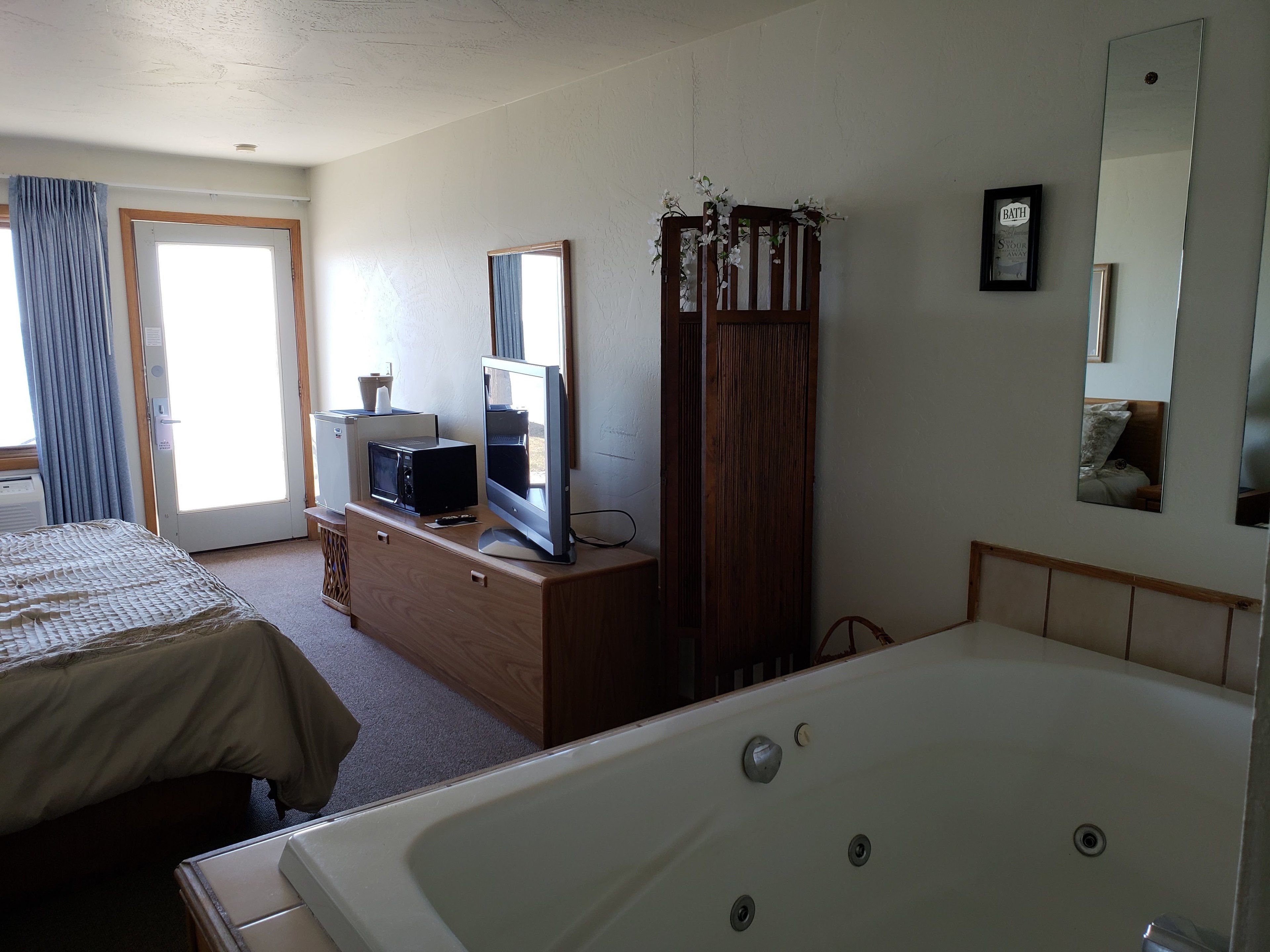 A hotel room with a large bed, a separate bathroom with a bathtub, a television on a wooden dresser, and a mirror on the wall.