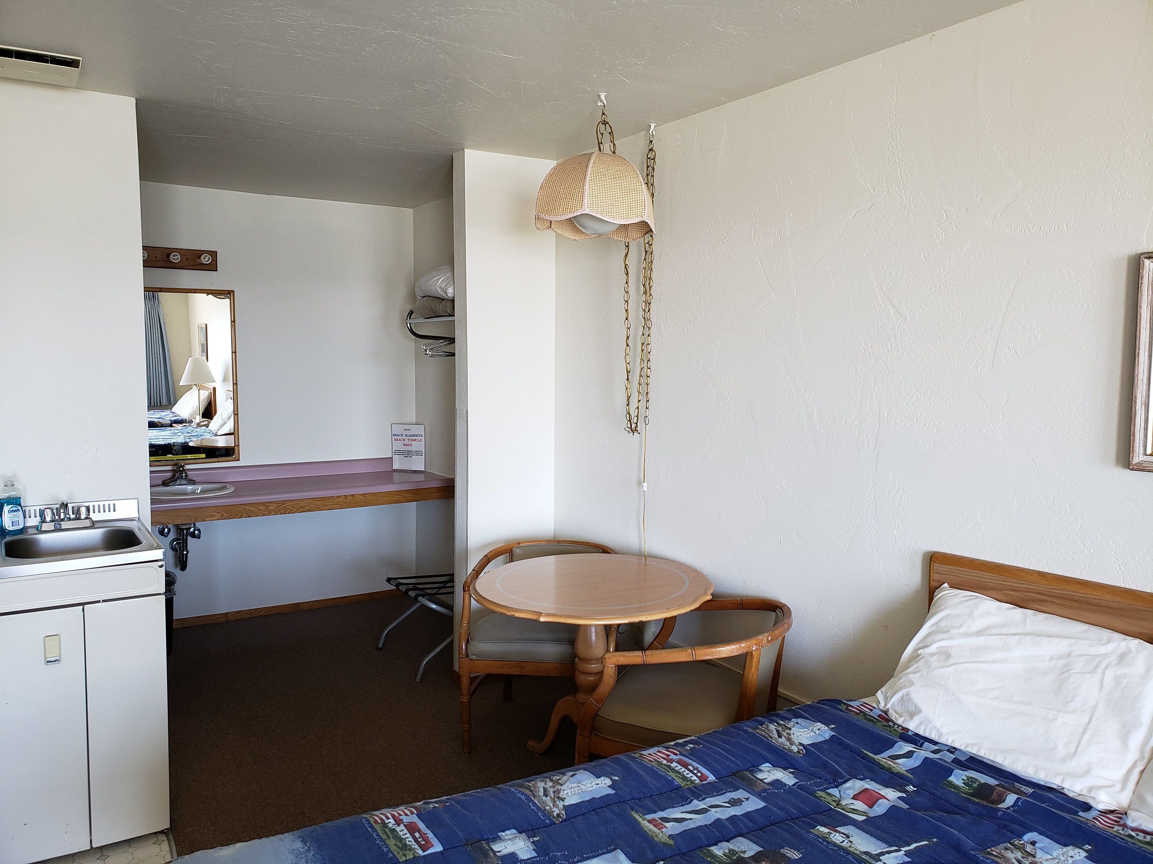A compact hotel room with a bed, a small desk with a sink, a chair, and a hanging light fixture.