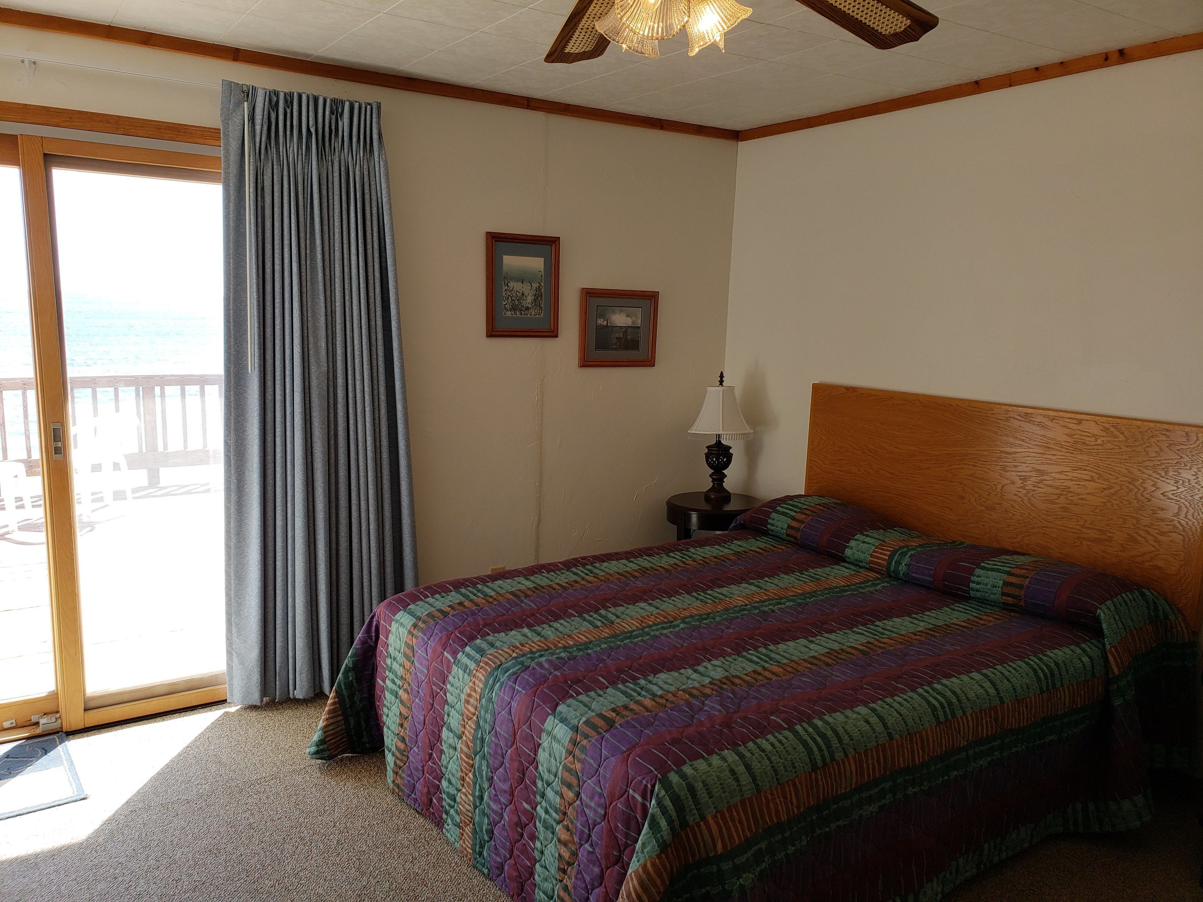 A hotel room with a large bed, a wooden headboard, a nightstand with a lamp, and a view of the ocean through a sliding door.