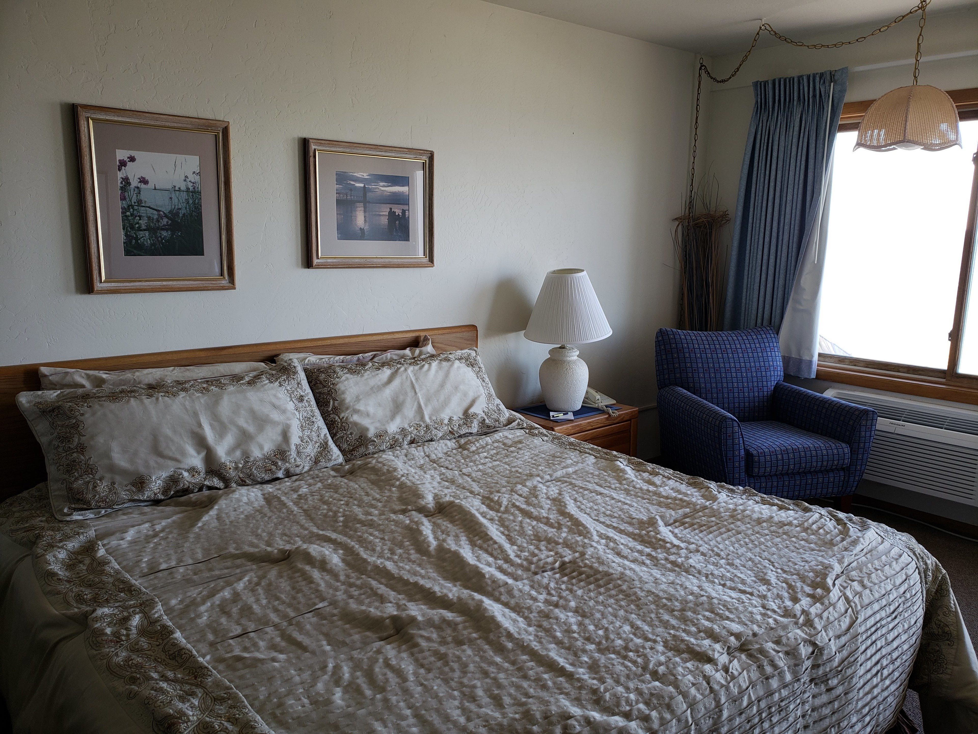 A neatly made bed with a patterned bedspread, a bedside lamp, two framed pictures on the wall, a blue chair, and a window with curtains.