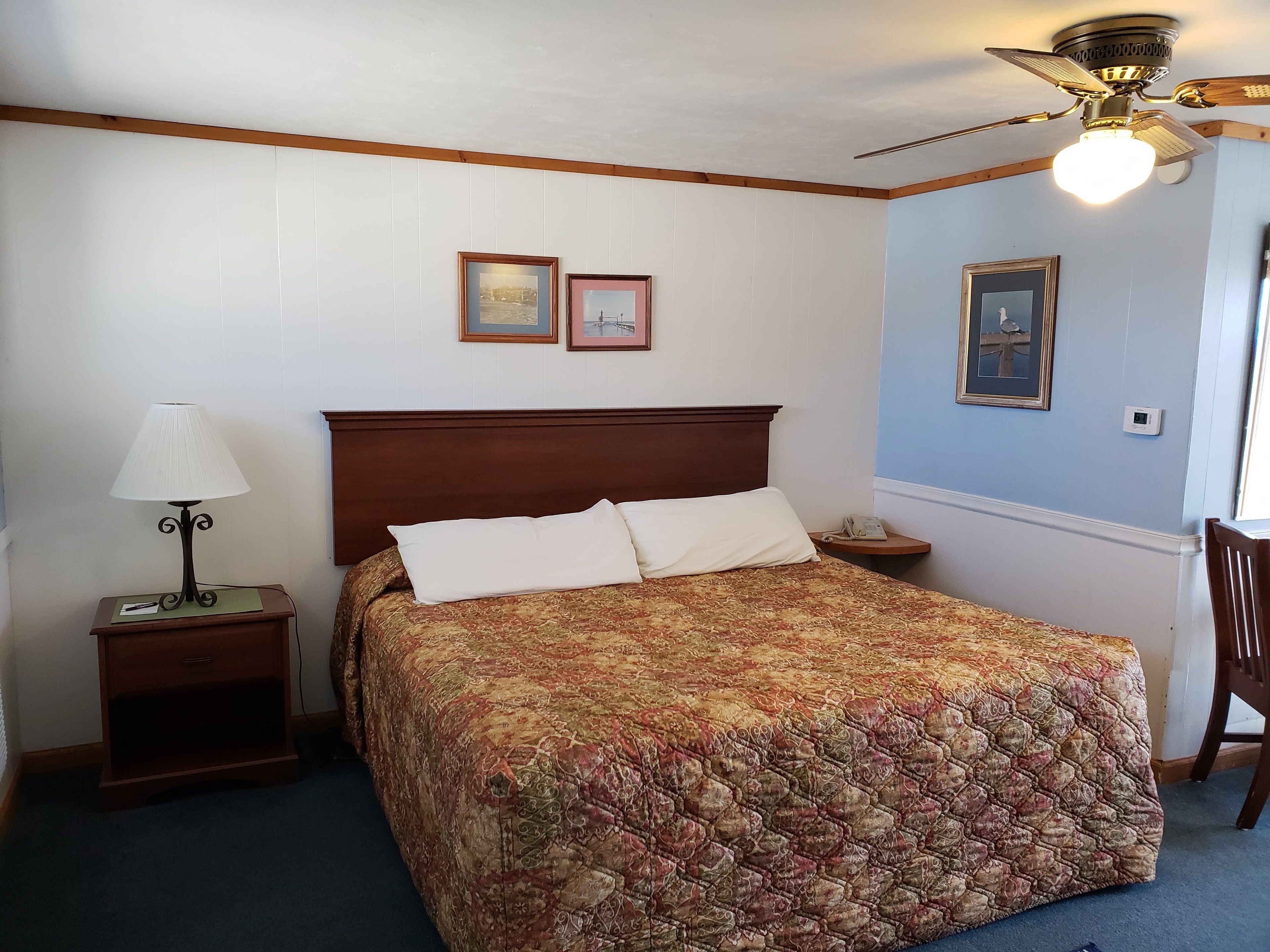 A neatly arranged bedroom with a wooden headboard, a bed with a patterned bedspread, a bedside table with a lamp, and framed pictures on the wall.