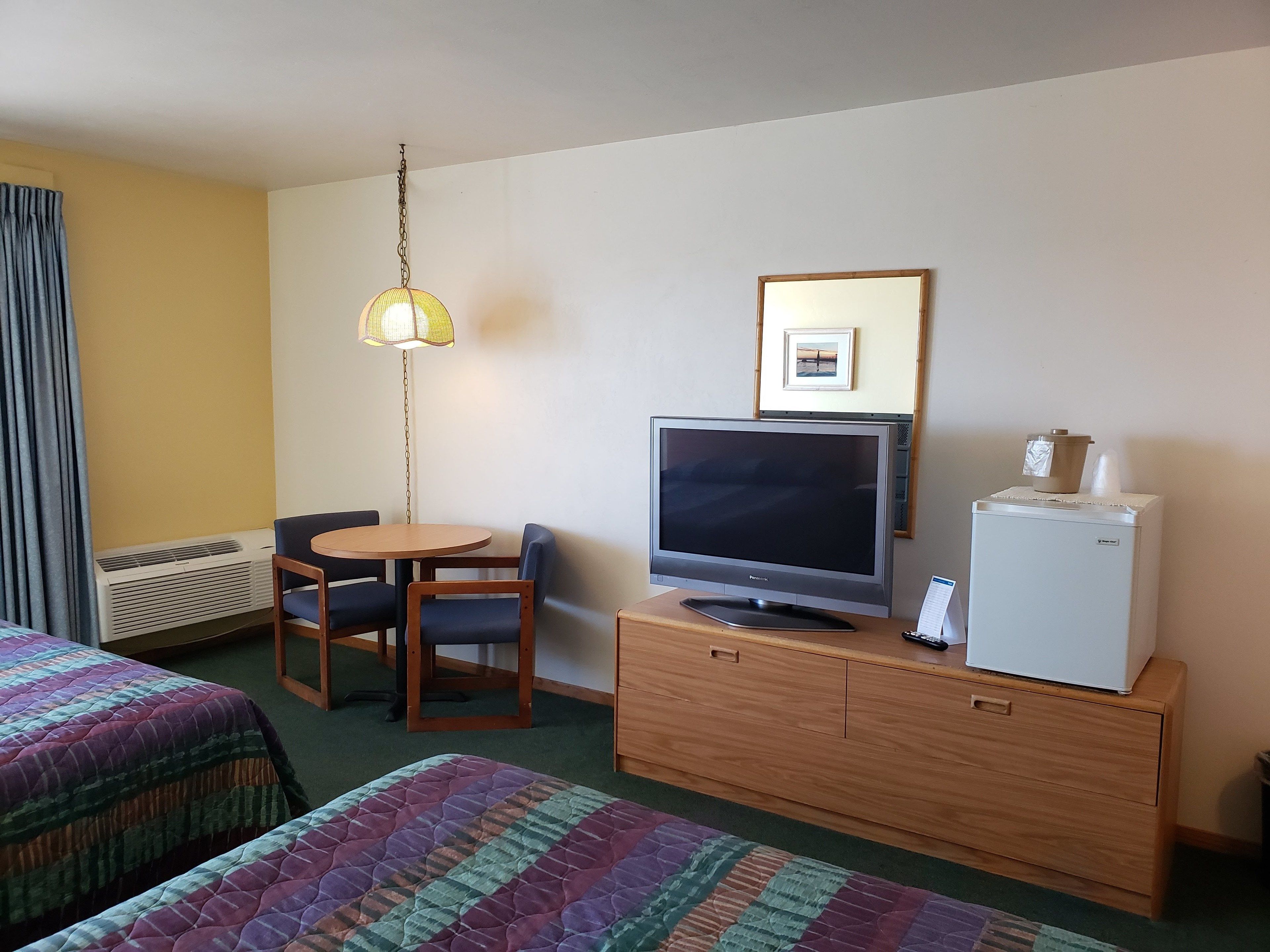 Une chambre d’hôtel équipée d’un lit, d’un bureau avec un ordinateur, d’une télévision, d’un réfrigérateur, d’une chaise, d’une table et d’une lampe suspendue.