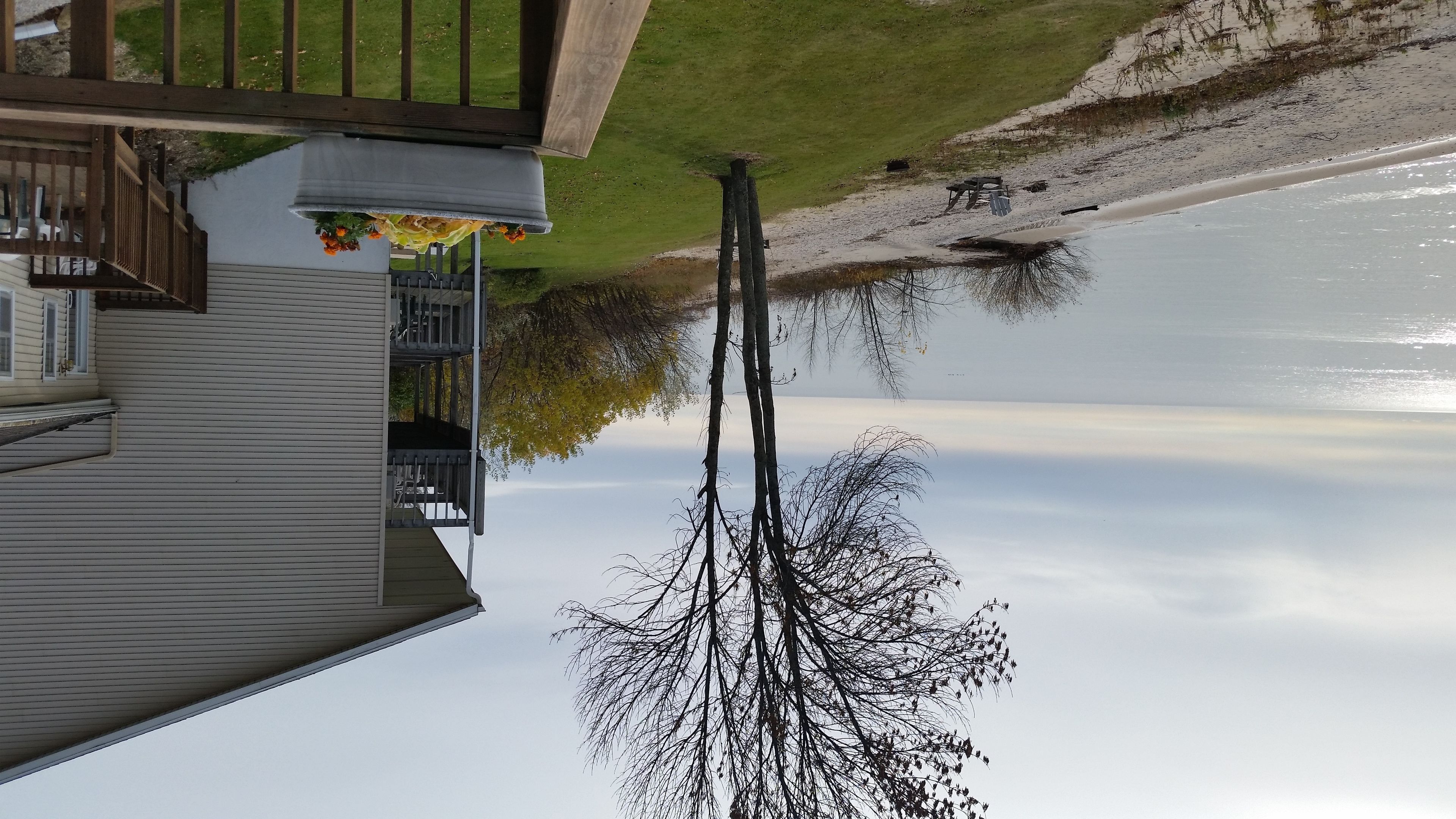 A house with a balcony overlooking a calm lake, a wooden deck, and a tree with bare branches.