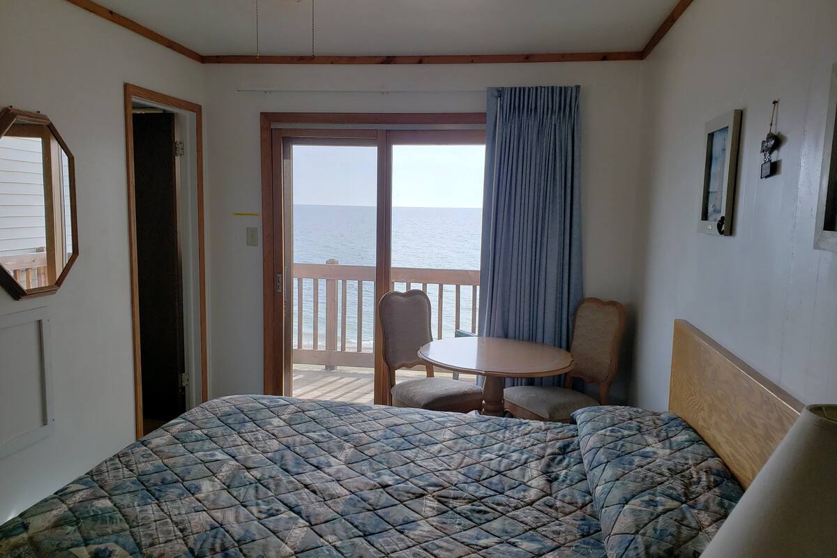 Un dormitorio con cama, una mesa y una silla. Se ve el océano a través de un ventanal corredero.