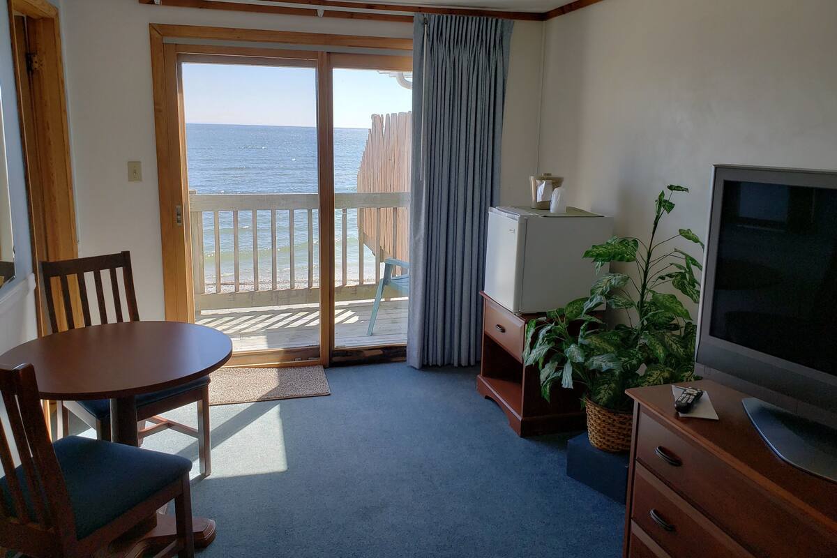 Habitación con puerta corredera de vidrio con marco de madera que da a un balcón con vista al mar. Hay una pequeña mesa con una silla, un televisor sobre un pedestal de madera y una planta en maceta.