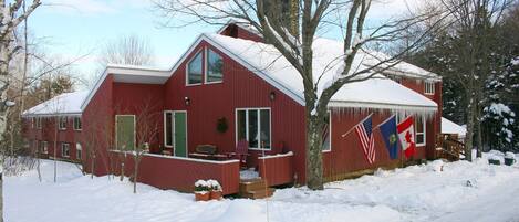 Une maison en bois rouge, avec un toit enneigé, un porche doté d’un banc et un drapeau sur le côté.