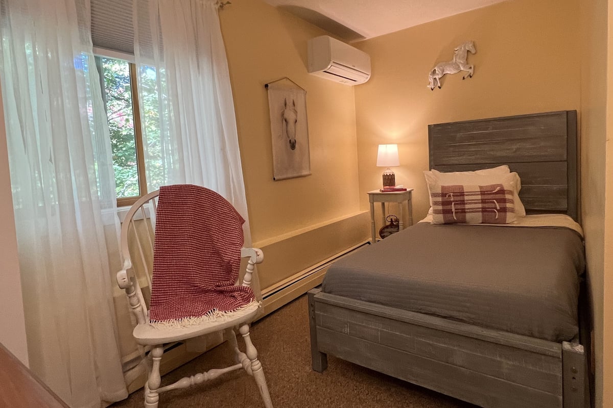 A bedroom with a bed, a chair, a nightstand with a lamp, an air conditioner, and a wall-mounted horse sculpture.