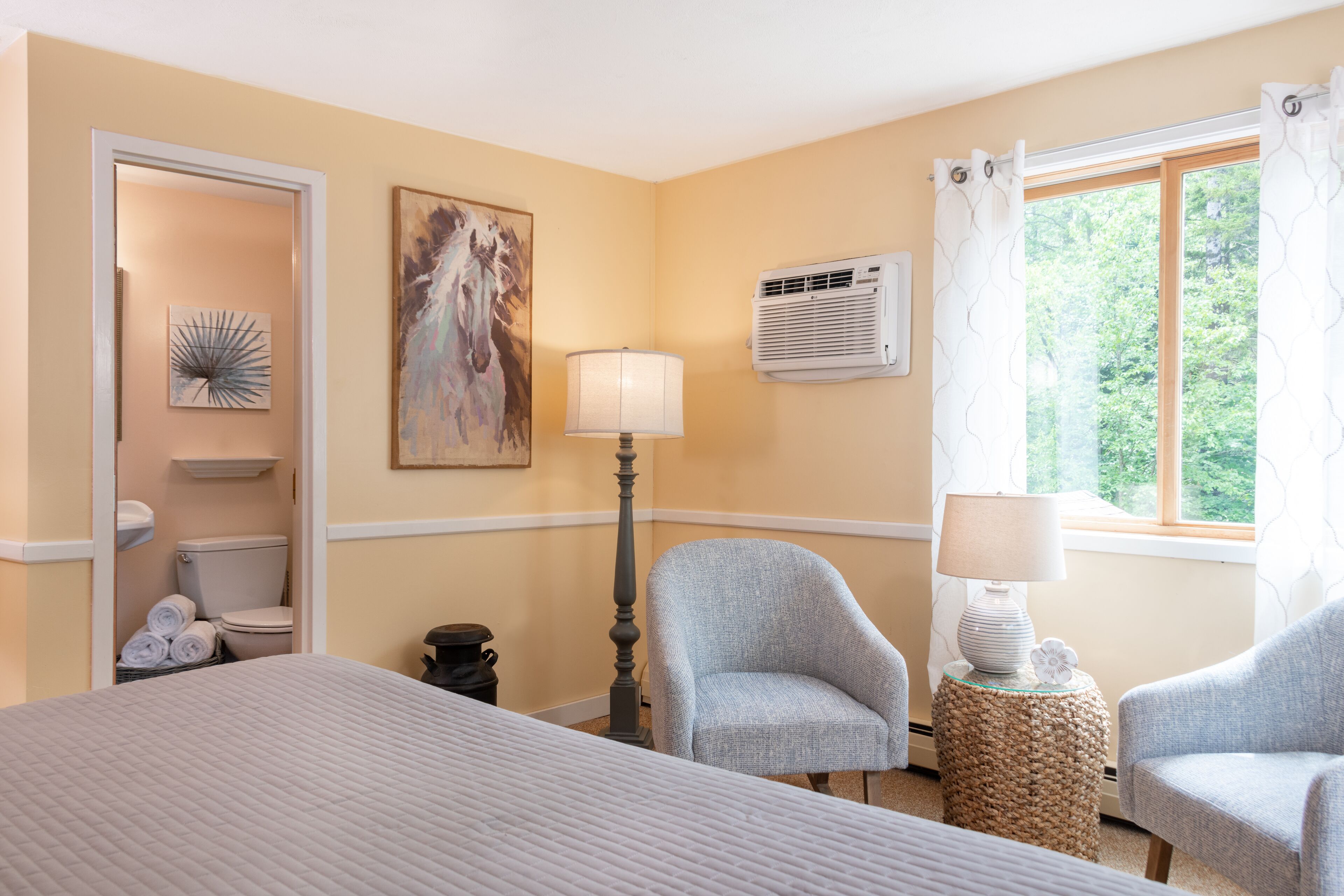 A hotel room with a bed, two armchairs, a lamp, a painting, an air conditioning unit, and a window with curtains.