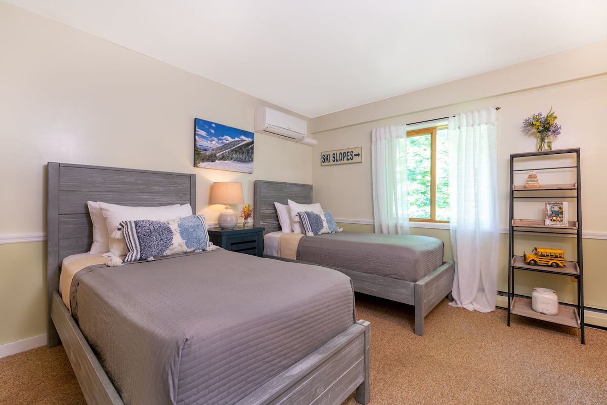 A bedroom with two beds, a nightstand, a shelf with toys, and a window with curtains.