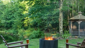 Ein Pavillon in bewaldeter Landschaft, in der Mitte ein Feuerkorb und zwei Adirondack-Stühle mit Getränken.