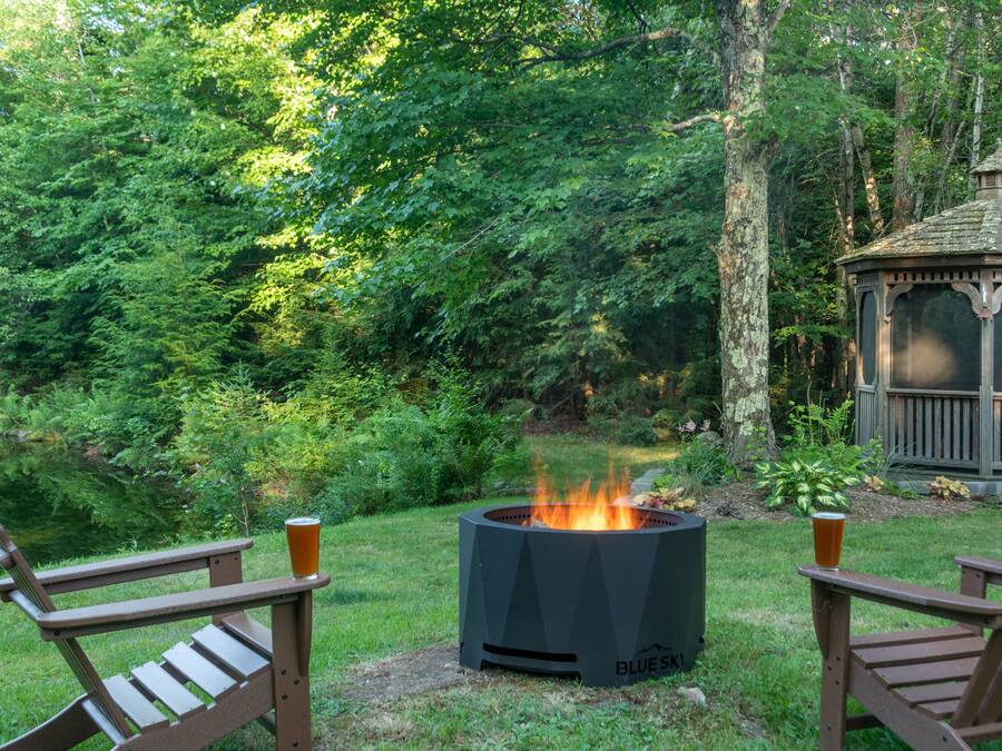 Un quiosco en un área boscosa con una chimenea en el centro y dos sillas Adirondack con bebidas.