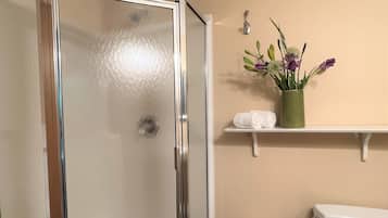 Et badeværelse med brusevæg i glas, toilet, håndvask, håndklædestang med håndklæder og en vase med blomster.