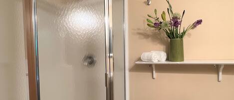 A bathroom with a glass shower door, a toilet, a sink, a towel rack with towels, and a vase with flowers.