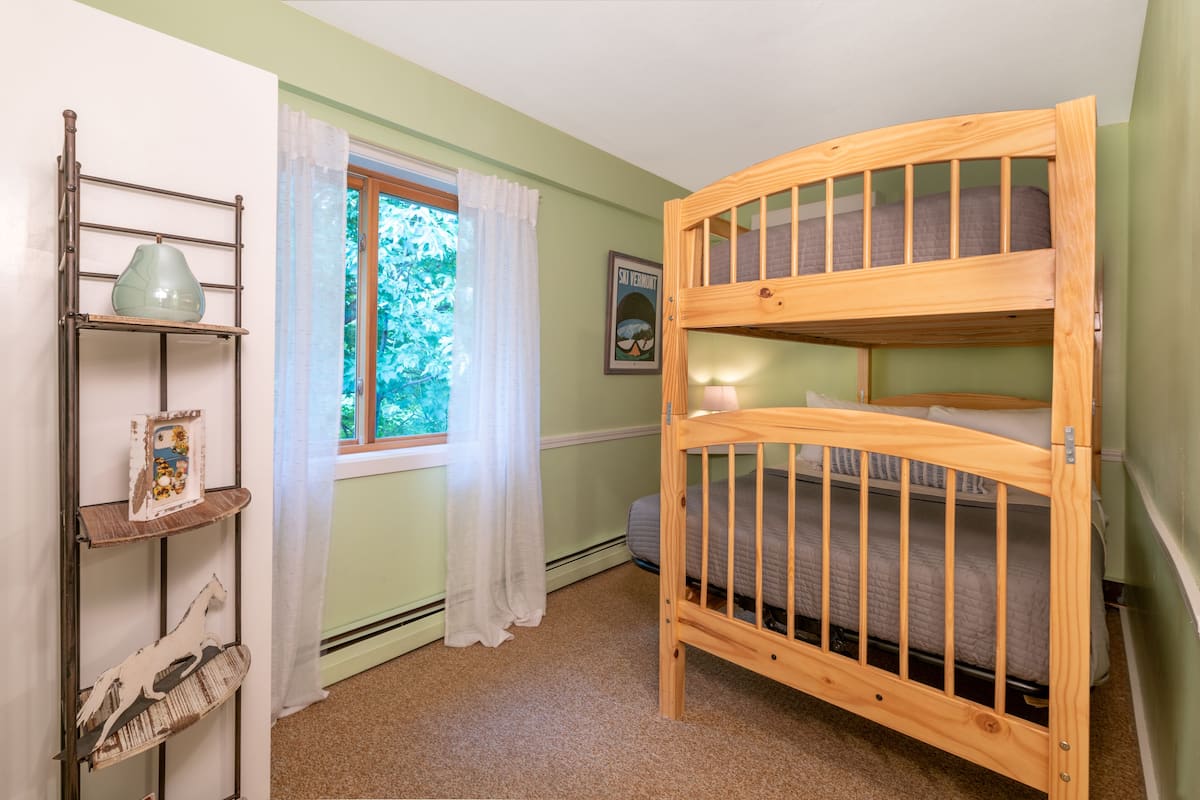 A child's bunk bed room with a window, a shelf with decorative items, and a framed picture on the wall.