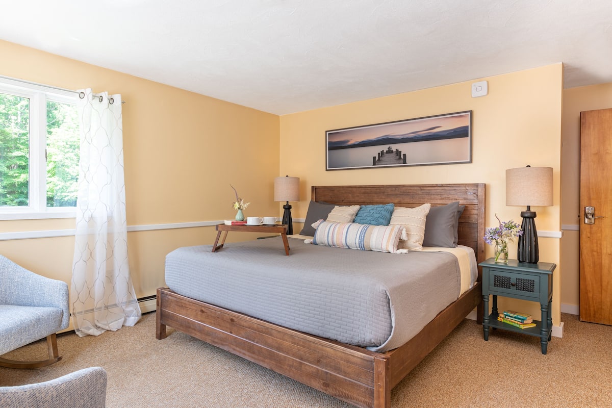 A bedroom with a wooden bed, a bedside table with a lamp, a chair, and a window with curtains.