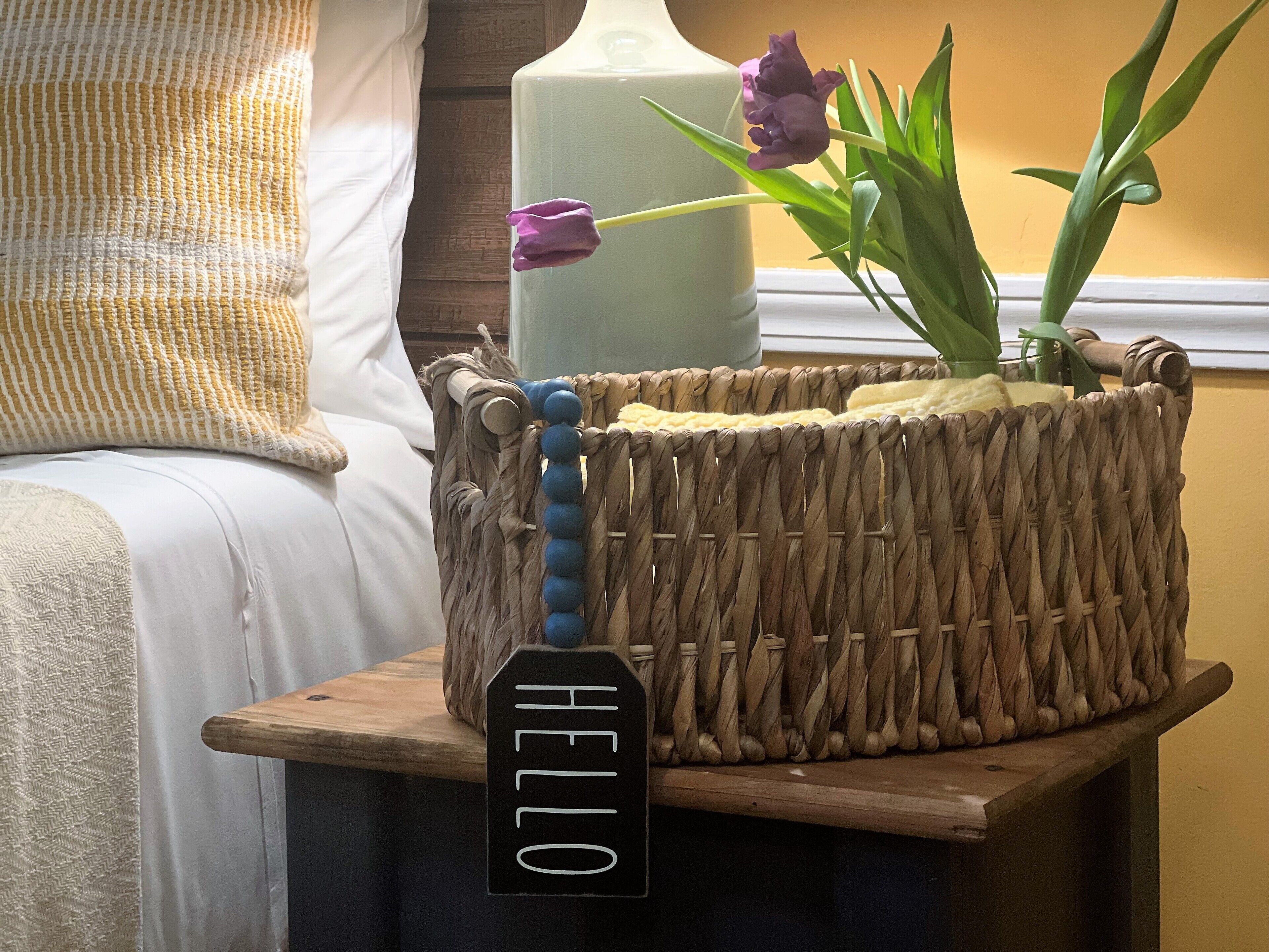 A woven basket with a wooden handle and a "HELLO" sign, placed on a wooden table next to a bed with a white pillow and a beige blanket.