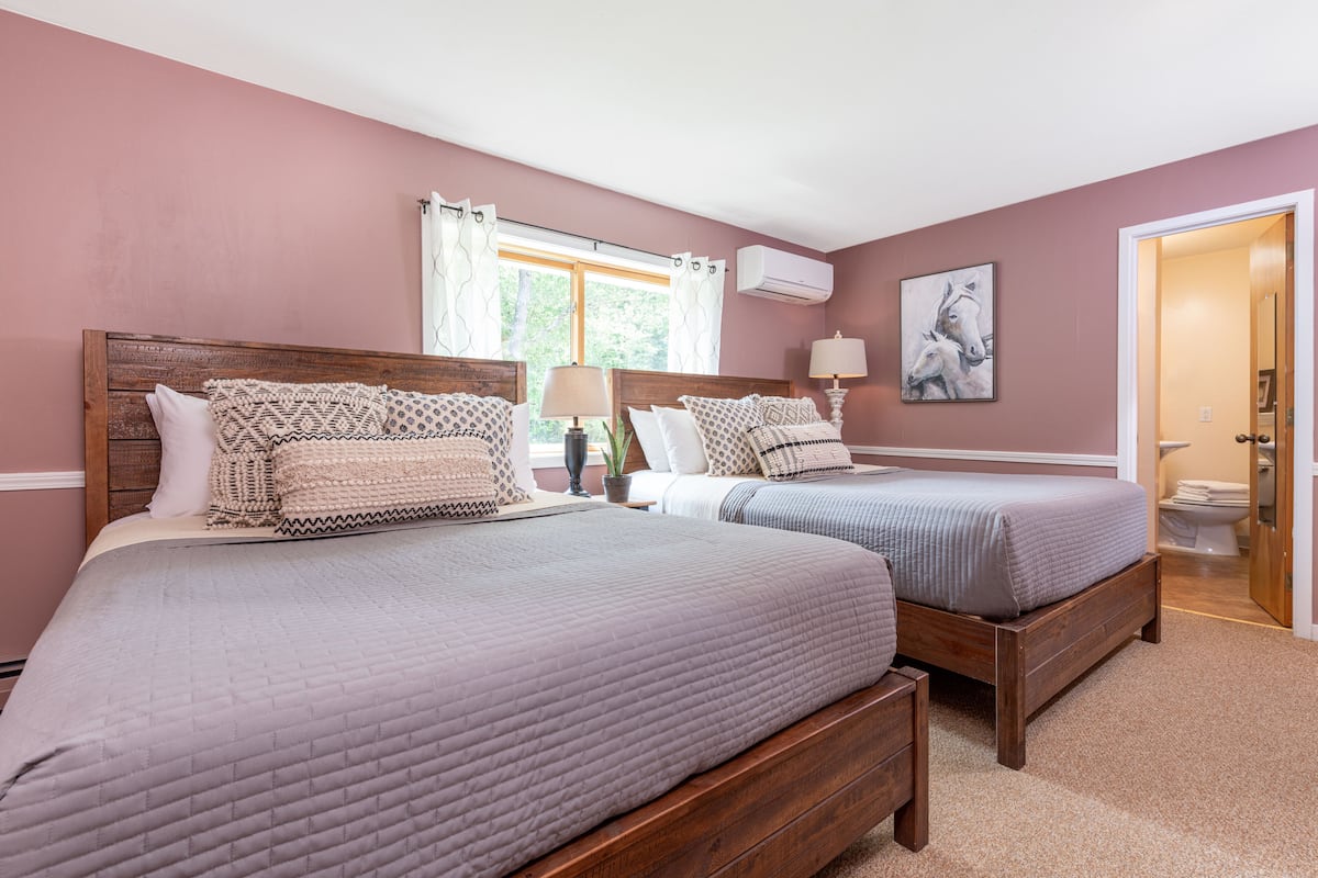A bedroom with two beds, a window with curtains, a wall-mounted air conditioner, and a painting.