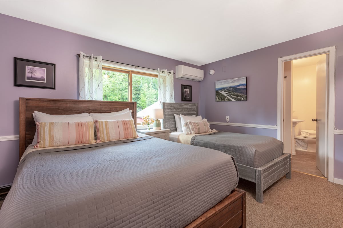 A bedroom with two beds, a window with curtains, a painting on the wall, and an air conditioning unit.