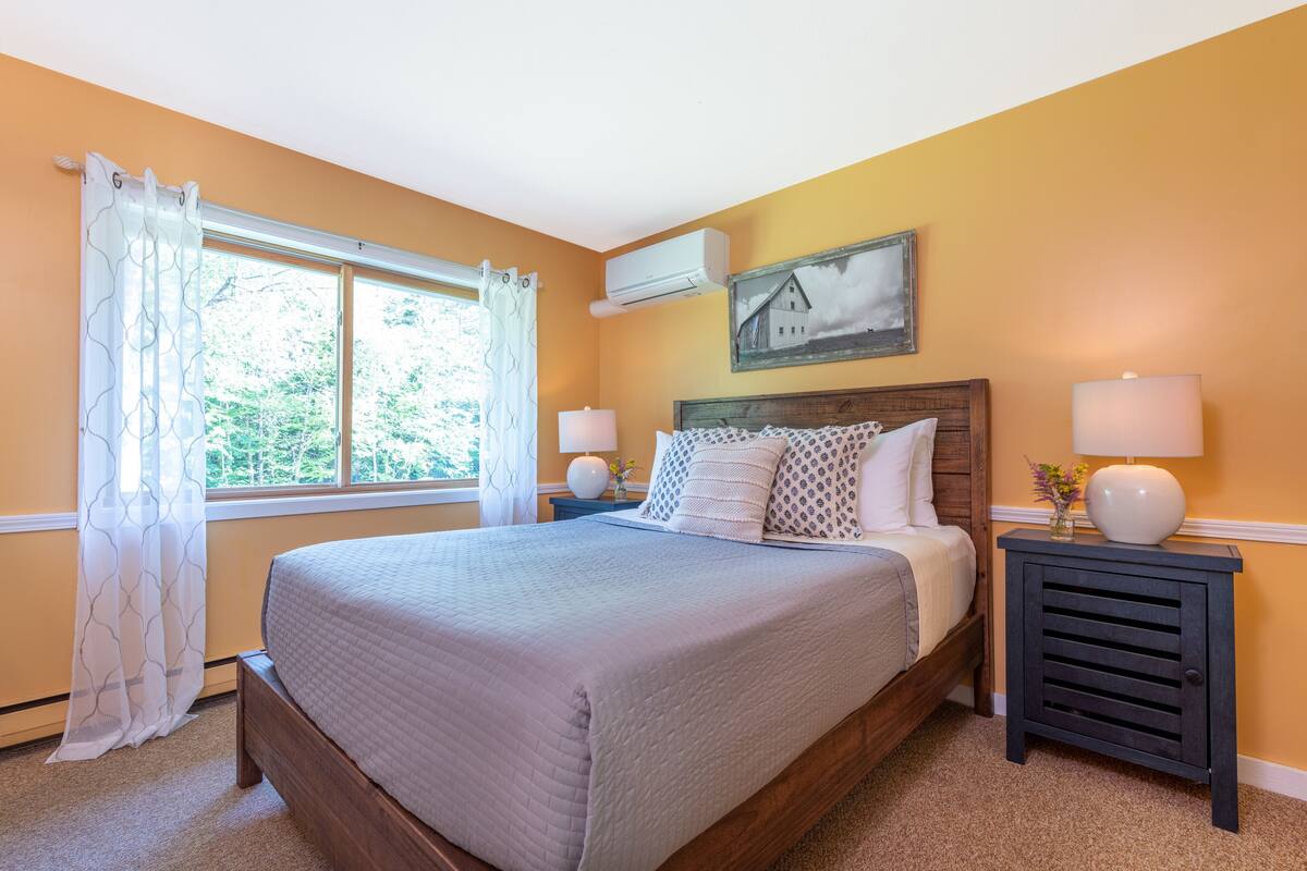 A bedroom with a large bed, two bedside tables, a window with curtains, an air conditioning unit, and a wall-mounted picture.