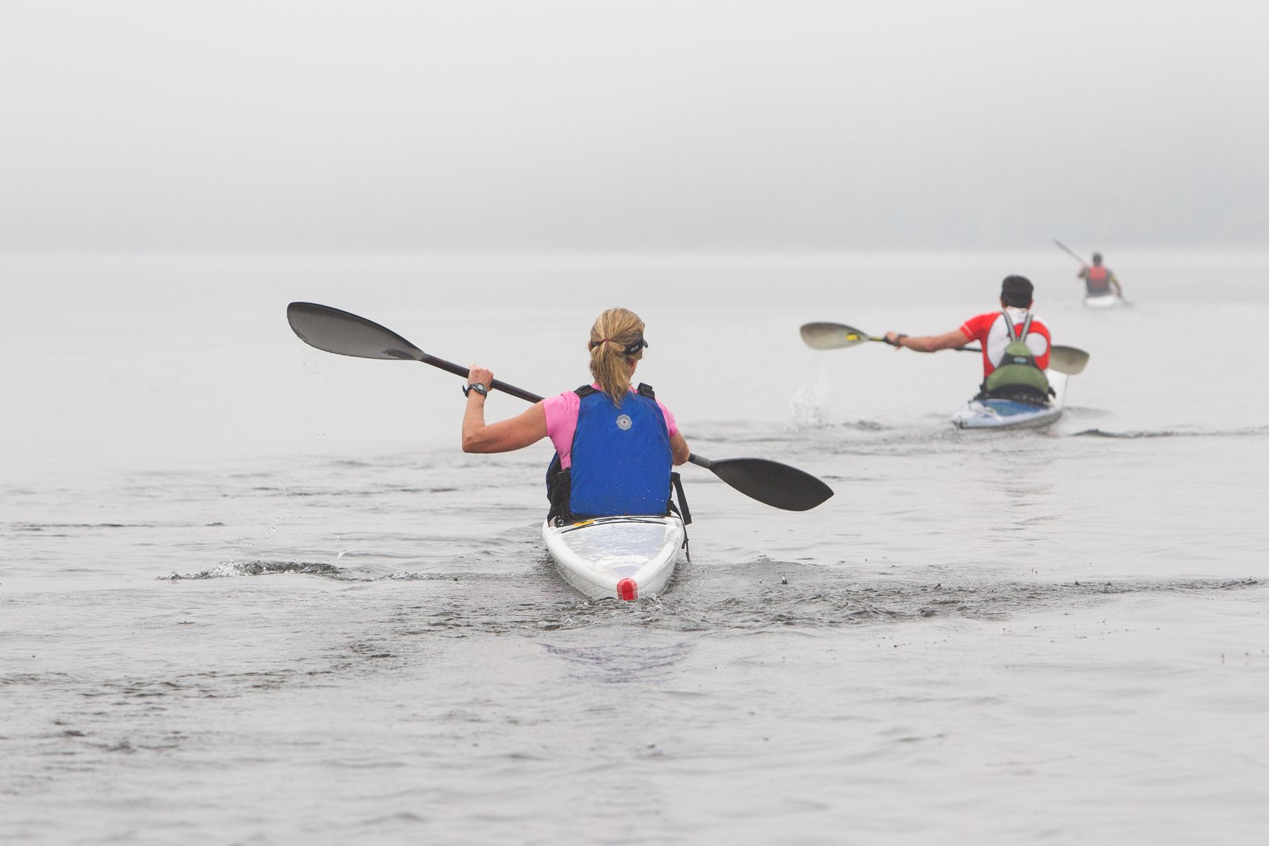 Un kayakista con chaleco salvavidas azul rema en un kayak blanco sobre una masa de agua brumosa.