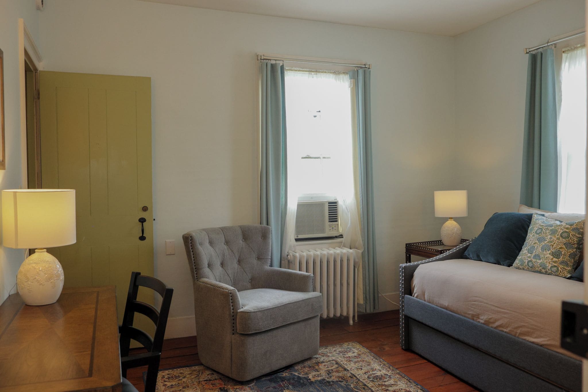 A hotel room with a bed, a chair, a desk, a radiator, and a window with curtains.