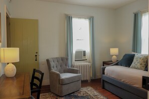 A hotel room with a bed, a chair, a desk, a radiator, and a window with curtains. - Centennial House (Northfield)