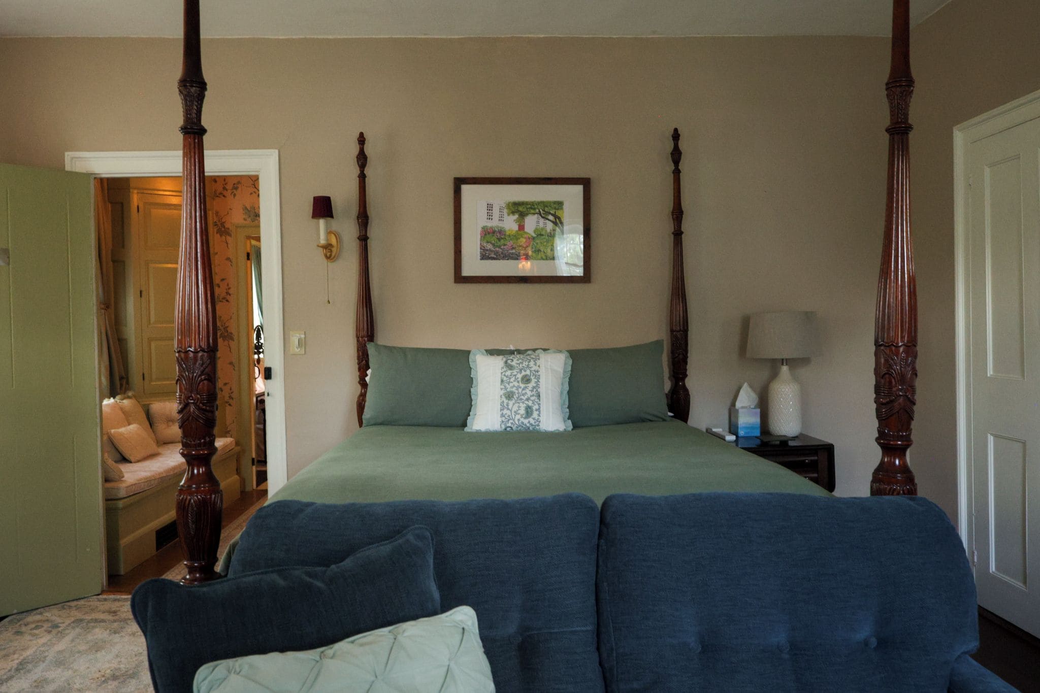 A four-poster bed with a green bedspread and a decorative pillow, a framed picture on the wall, and a bedside table with a lamp.