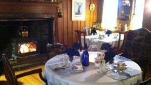 A dining area with a round table set for four, surrounded by wooden chairs. A fireplace is visible in the background, and the room has wooden paneling. - Centennial House (Northfield)
