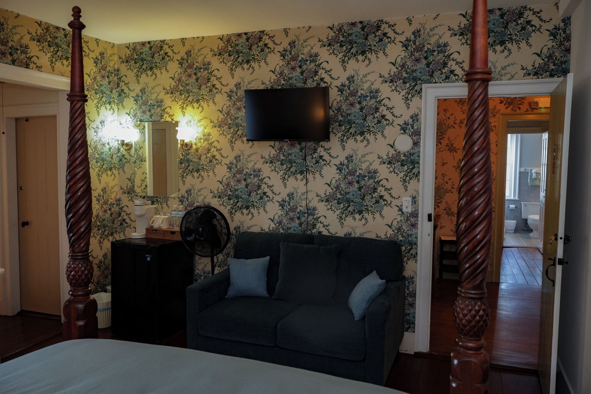 Un dormitorio con una cama con dosel, un televisor de pantalla plana montado en la pared, un sofá de color oscuro y un aparador de madera.