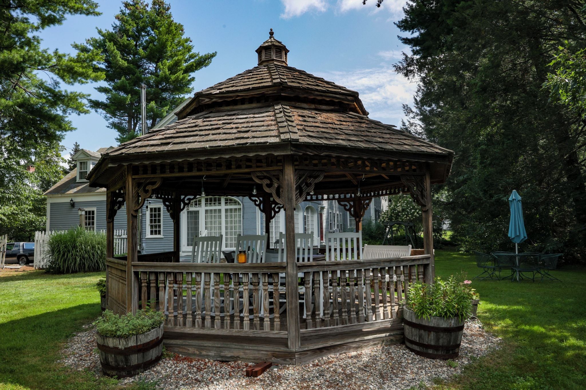 A wooden gazebo with a peaked roof, surrounded by a gravel area with potted plants.