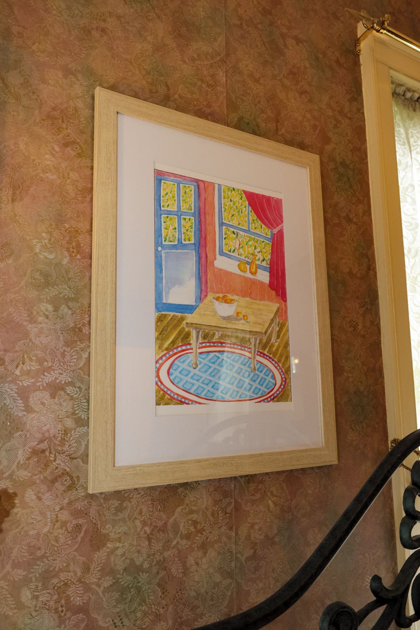A framed artwork depicting a room with a window, a table with fruit, and a patterned rug.