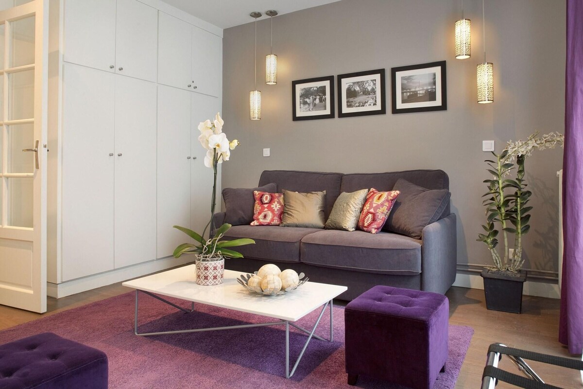 Een moderne woonkamer met een grijze bank, een witte salontafel, een paars voetenbankje en een potplant.