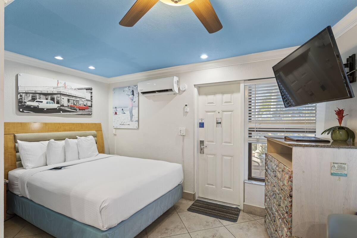 A hotel room with a bed, a ceiling fan, an air conditioning unit, a television, a door, and a window with blinds.