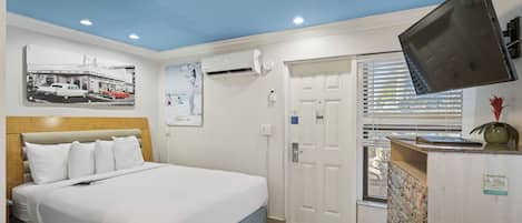 A hotel room with a bed, a ceiling fan, an air conditioning unit, a television, a door, and a window with blinds.