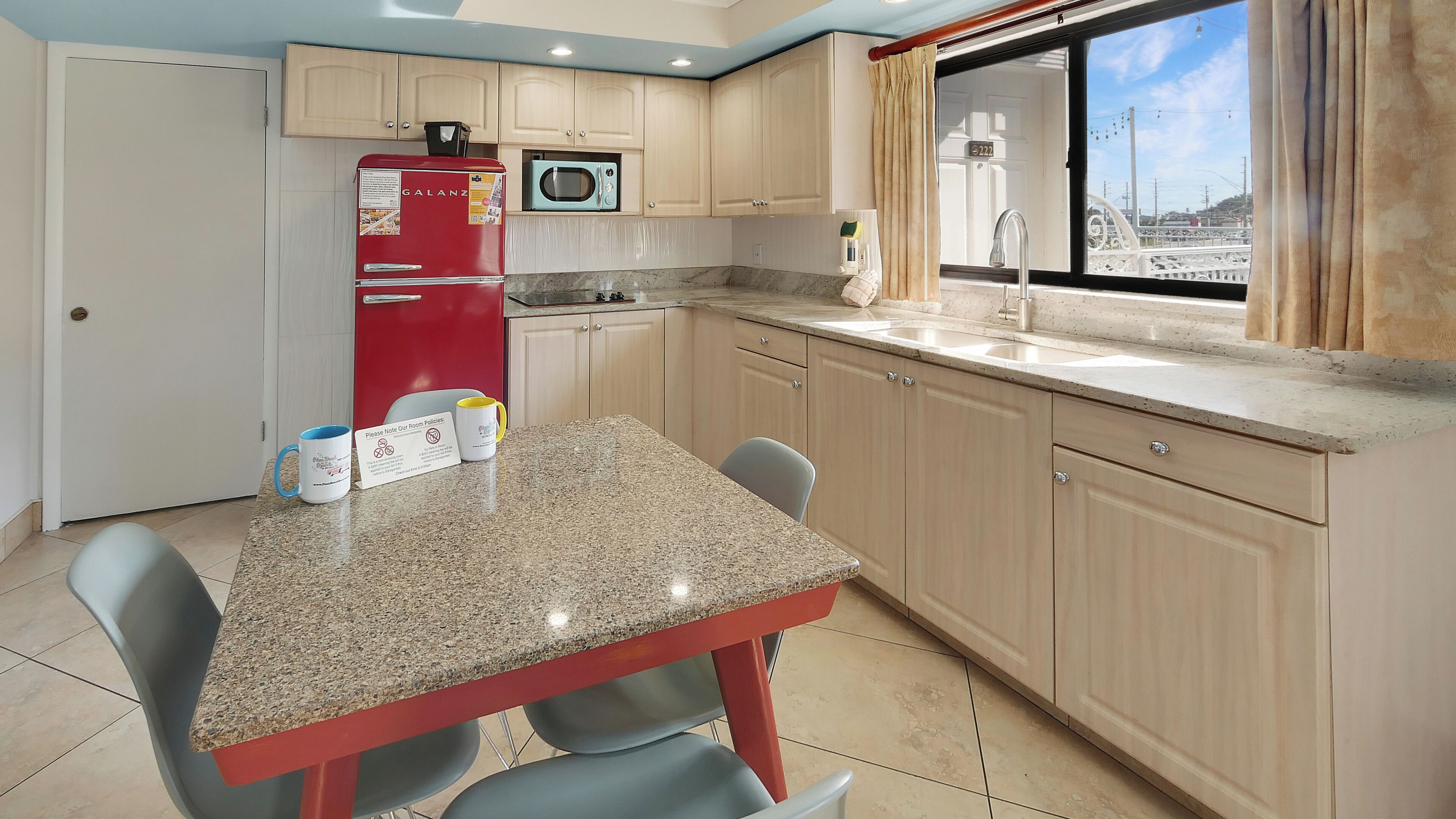A kitchen with a granite countertop, a red refrigerator, a microwave, and a window with curtains.