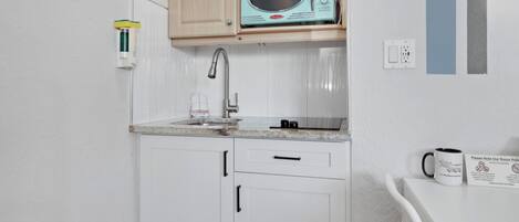 A kitchenette with a microwave, a faucet, and a sink. There is a cabinet with a door and a countertop with a coffee mug.