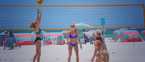 A beach volleyball game with players in swimwear, a volleyball net, and beach umbrellas in the background.