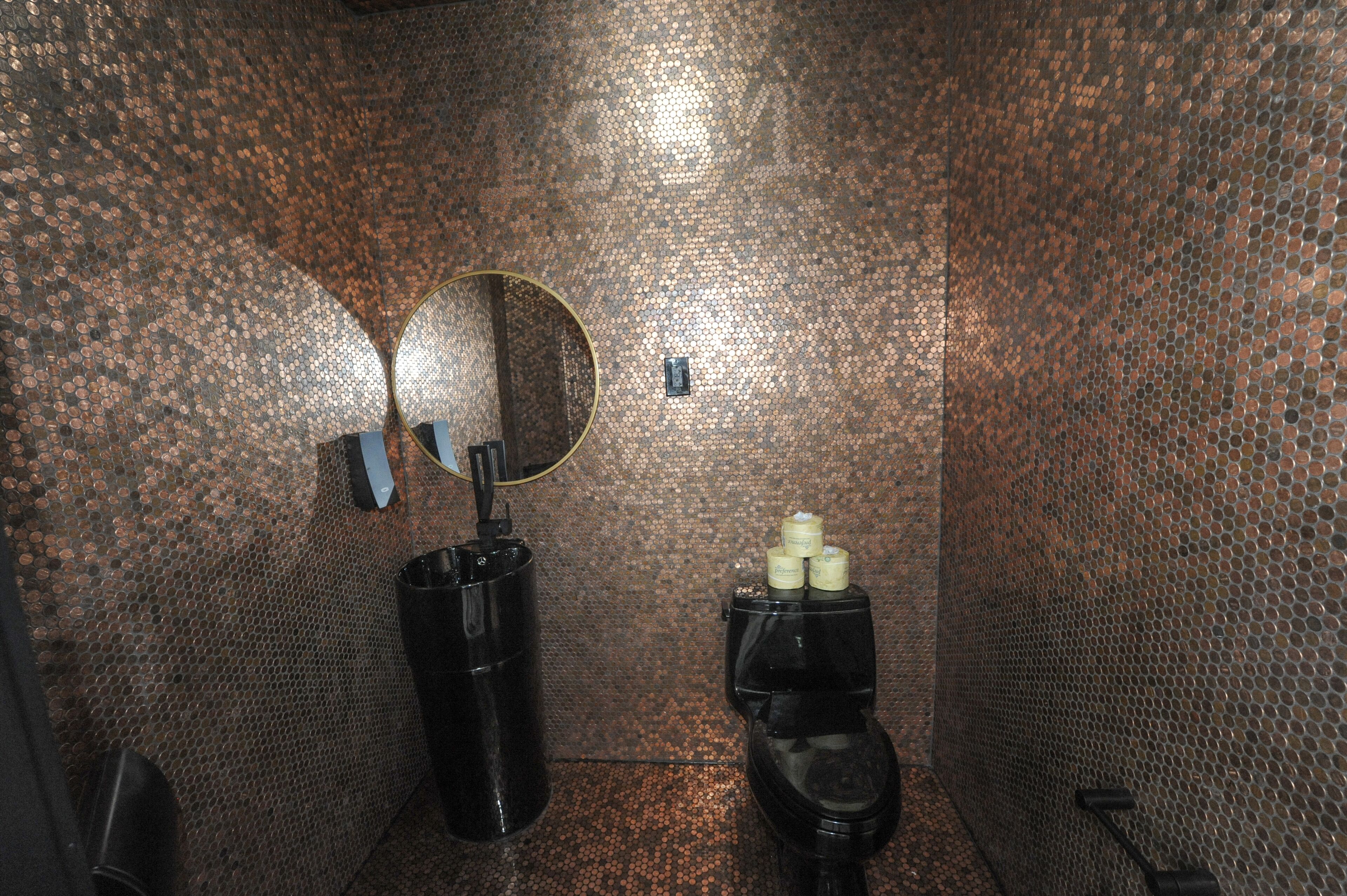 A restroom with a copper-colored tiled wall, a round mirror, a black trash bin, and a toilet with a small trash can beside it.