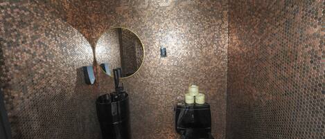 A restroom with a copper-colored tiled wall, a round mirror, a black trash bin, and a toilet with a small trash can beside it.