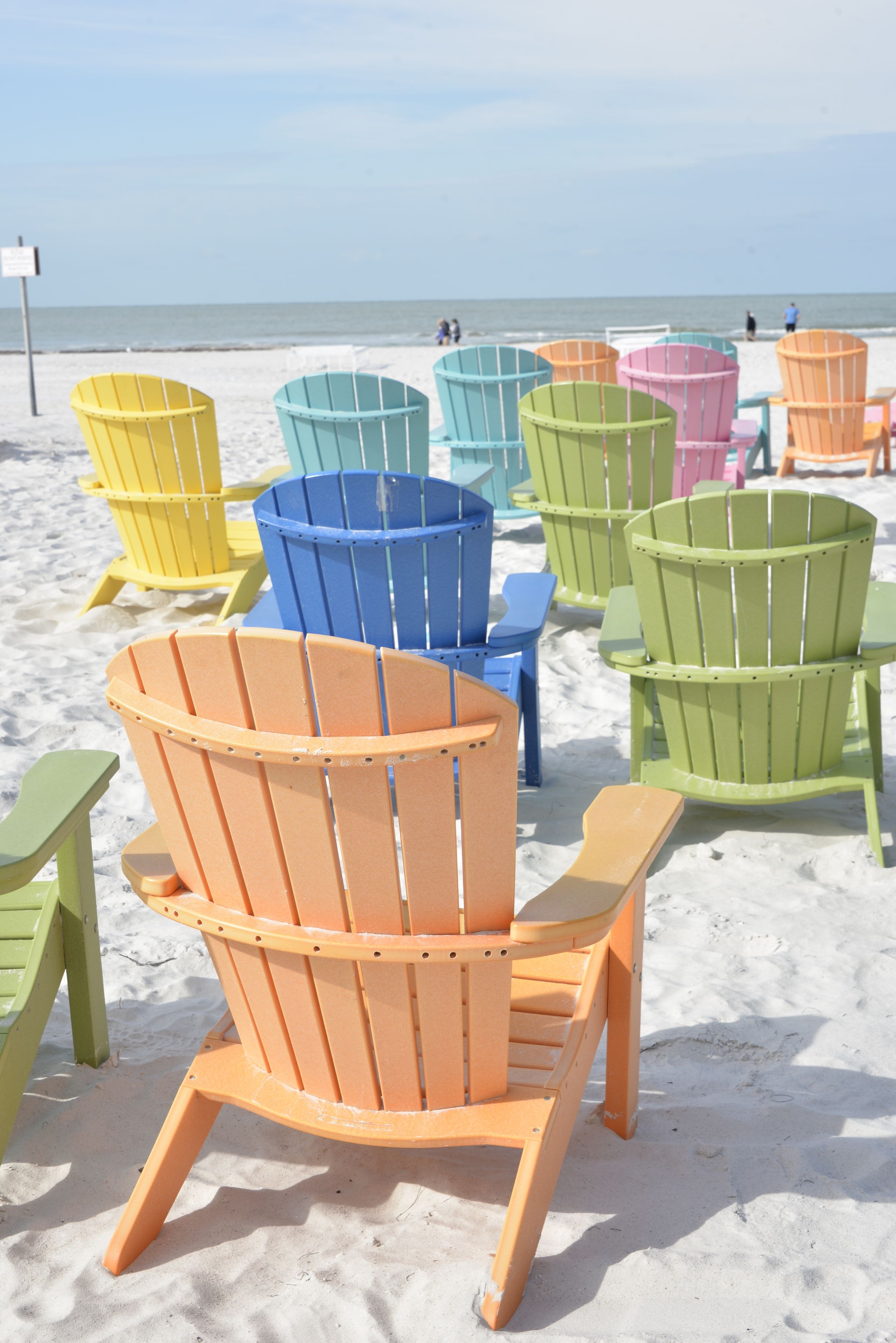 Une rangée de chaises Adirondack aux couleurs vives, sur une plage de sable, avec l’océan en arrière-plan.