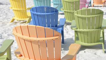 Eine Reihe bunter Adirondack-Stühle an einem sandigen Strand, im Hintergrund das Meer.