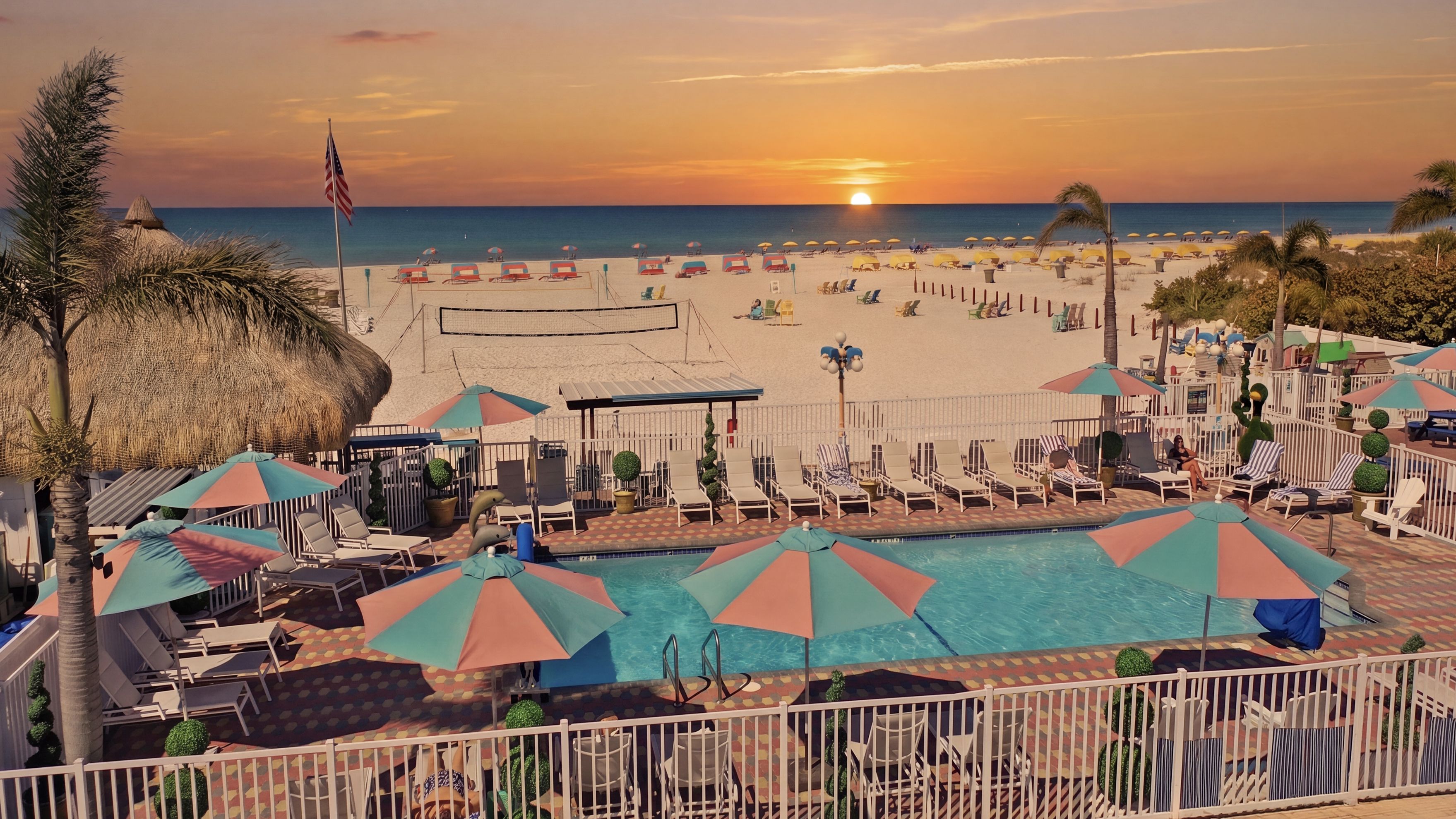 A beach resort with a swimming pool, sun loungers, umbrellas, and a view of the sunset over the ocean.