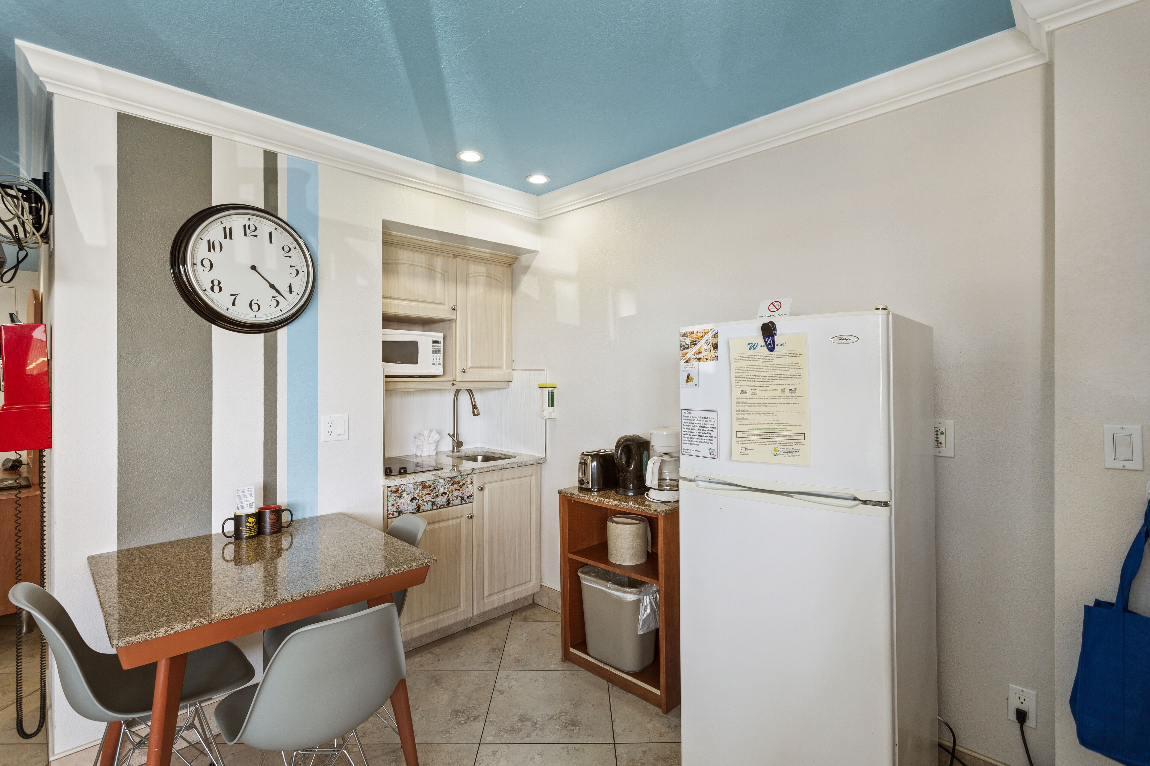Una cocina compacta con un reloj, microondas, refrigerador y un comedor con sillas.