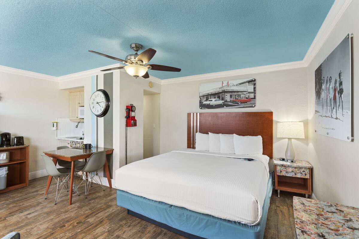 A hotel room with a bed, a desk, a chair, a ceiling fan, a clock, a fire extinguisher, and a framed picture on the wall.