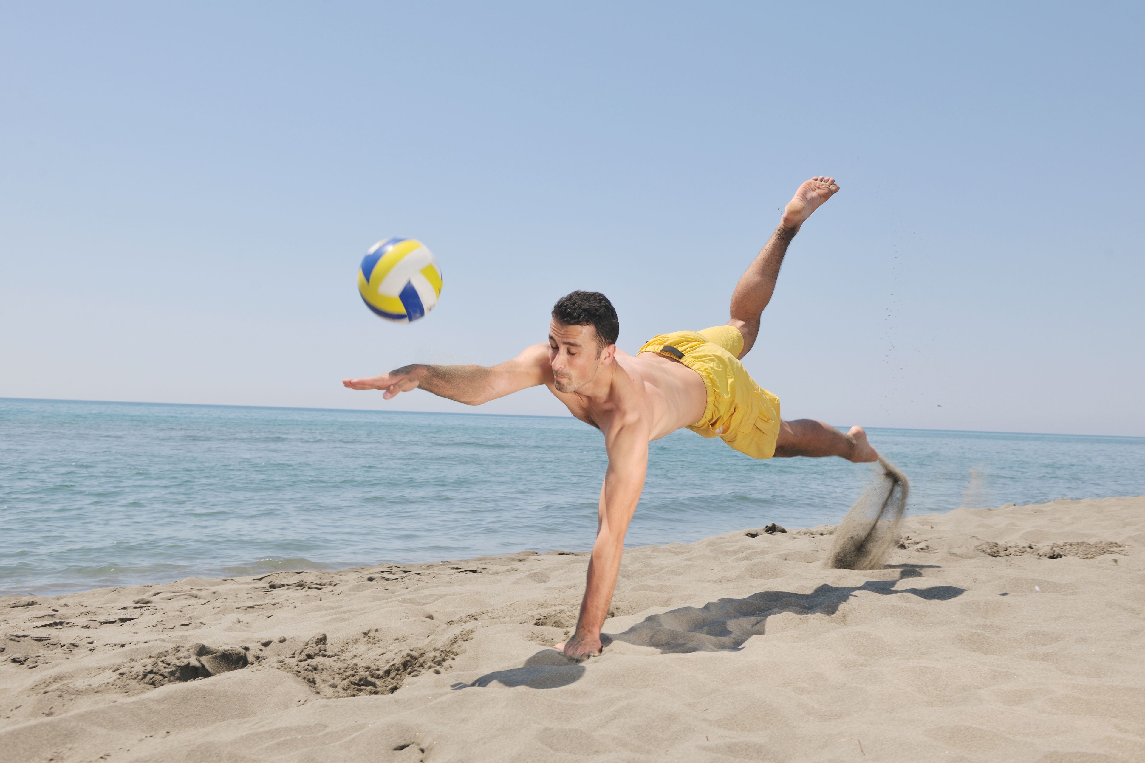A person performing a diving stunt on a sandy beach with a clear sky and calm sea in the background.
