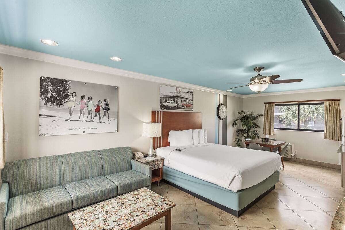 A hotel room with a bed, a sofa, a small table, a nightstand, a ceiling fan, a window with curtains, and a wall clock.