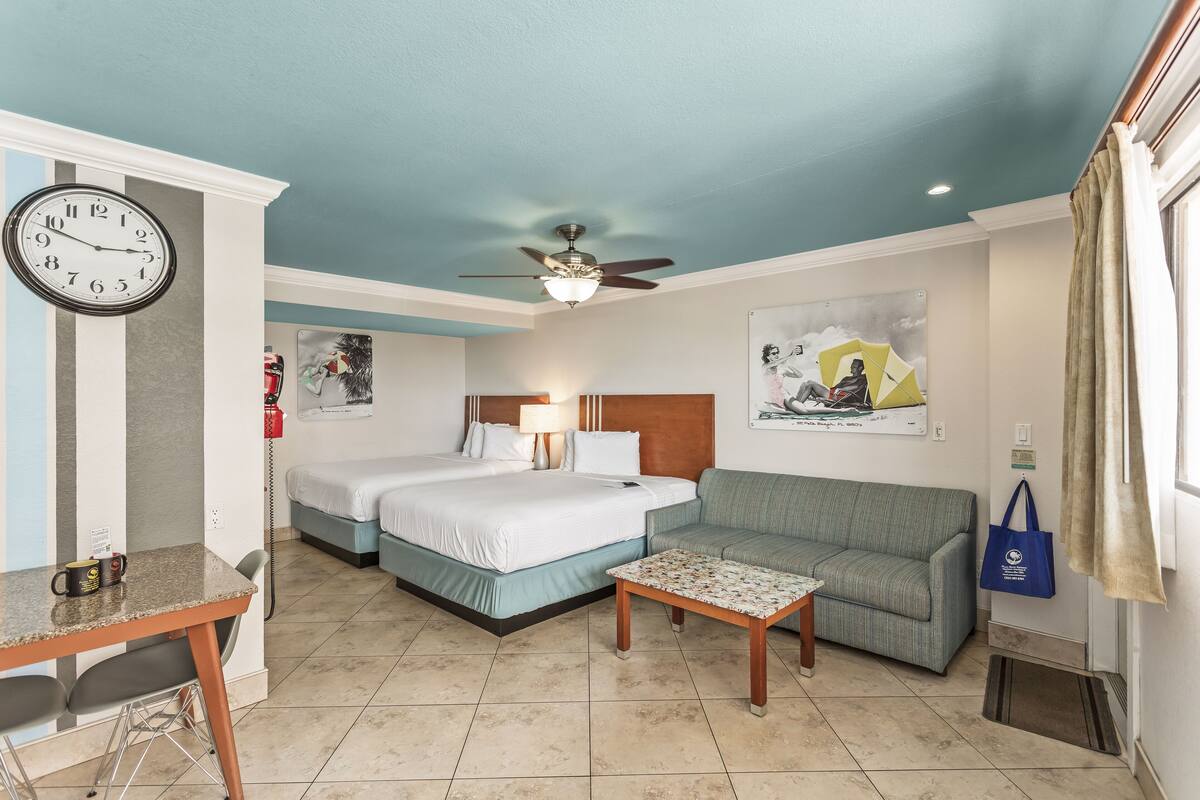 A hotel room with two beds, a sofa, a coffee table, a clock, and a ceiling fan.